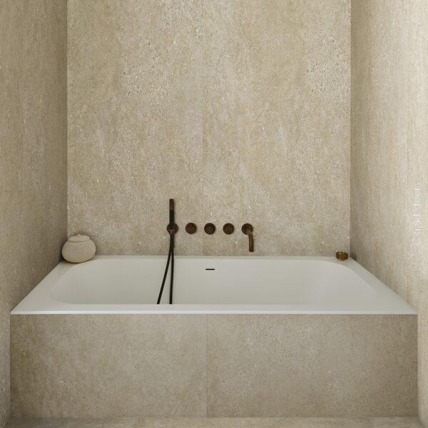Baignoire blanche encastrée entourée de carrelage en pierre beige. Robinetterie murale minimaliste en bronze. Ambiance spa.