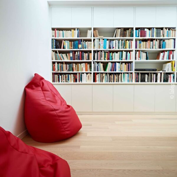 Espace lecture minimaliste avec bibliothèque blanche encastrée et parquet clair. Deux poufs rouges vif; un livre affiche « Atlas Ferraris »