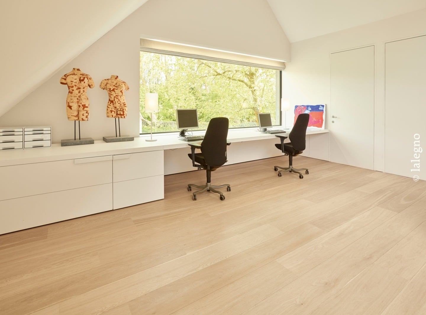 Bureau minimaliste aménagé sous les combles. Long plan de travail blanc pour deux postes, grande fenêtre donnant sur les arbres. Plancher bois clair.