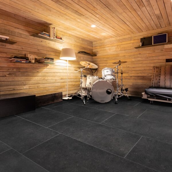 Salle de musique rustique aux murs et plafond en bois clair. Carrelage rectangulaire noir au sol, batterie blanche, lampadaire et livres.