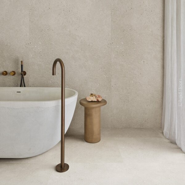 Salle de bain minimaliste. Carrelage beige effet pierre sur les murs et le sol. Baignoire, robinetterie bronze et rideaux blancs.