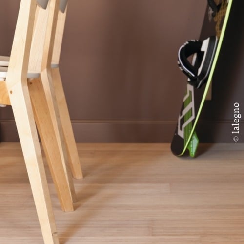 Pieds de chaise en bois clair et snowboard noir et vert design contre un mur marron. Le sol est en bois naturel.