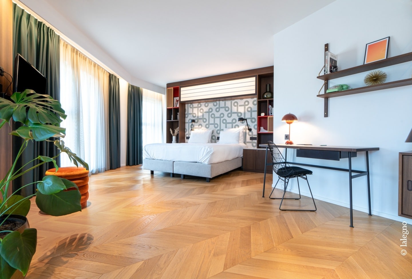 Suite d'hôtel design: parquet chevron, lit double, étagères en bois sombre, bureau épuré, lampe orange et rideaux verts.