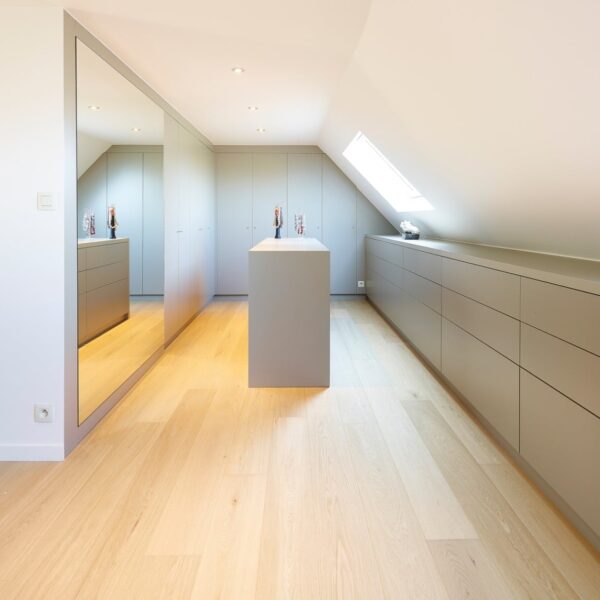 Dressing sous-pente moderne avec armoires, îlot central, et long meuble à tiroirs gris clair. Grand miroir mural et sol en chêne.