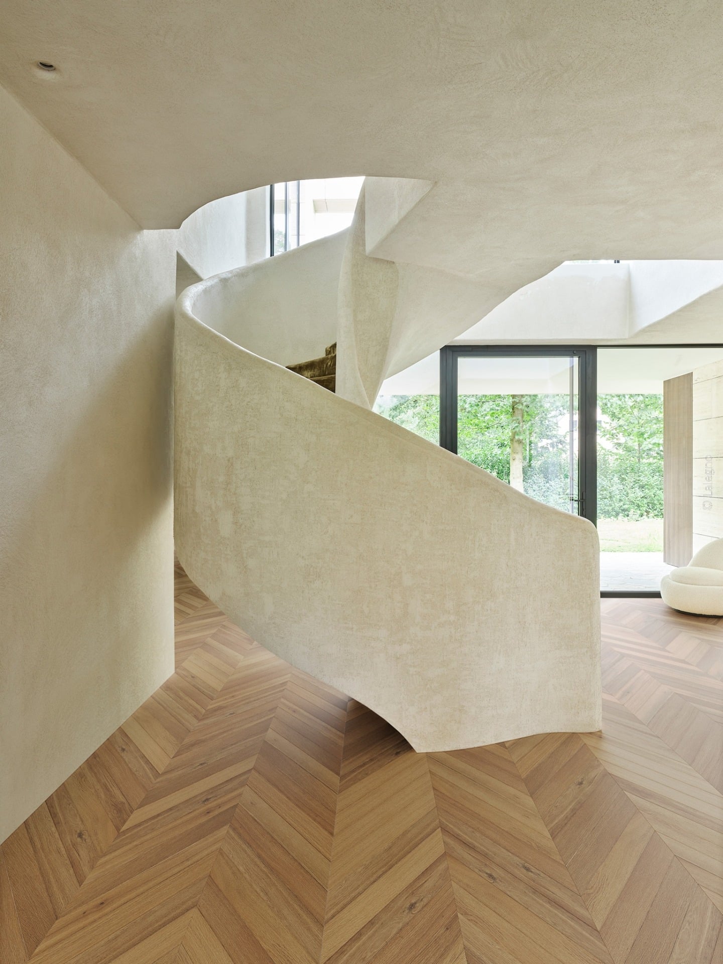 Escalier courbe sculptural en stuc beige texturé dans un hall minimaliste. Parquet chevron bois. Lumière naturelle par porte-fenêtre.