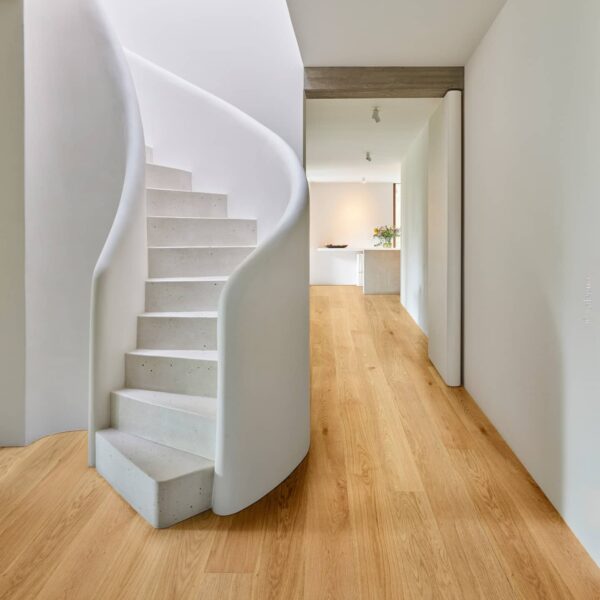 Couloir minimaliste avec escalier courbe blanc et marches en béton. Sol en parquet de bois clair. Vue sur la cuisine.