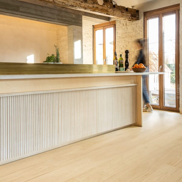 Îlot de cuisine lumineuse en bois clair avec panneau cannelé et laiton. Parquet chêne moderne. Poutres apparentes rustiques en hauteur.