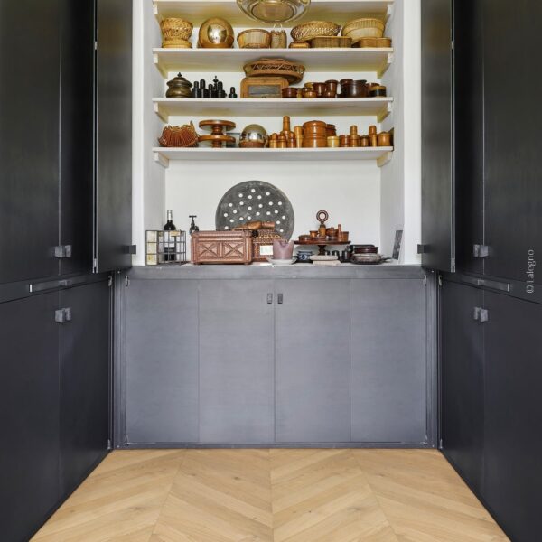 Niche de rangement moderne avec étagères blanches exposant des objets rustiques en bois et osier. Placards noirs et parquet chevron.