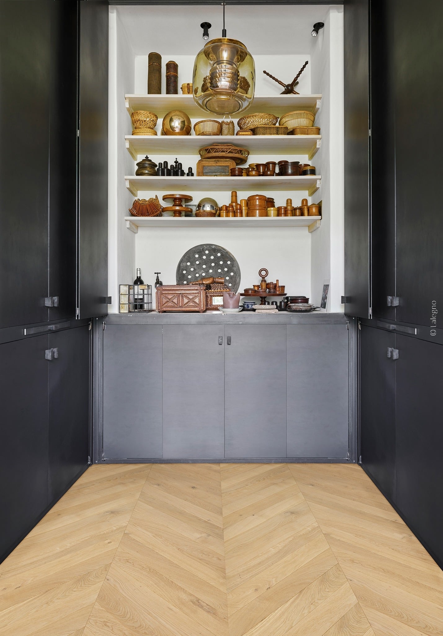 Niche de rangement moderne avec étagères blanches exposant des objets rustiques en bois et osier. Placards noirs et parquet chevron.