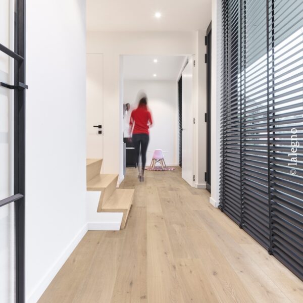 Couloir design avec parquet chêne clair et escalier moderne. Stores vénitiens sombres. Silhouette floue marchant vers une pièce lumineuse.