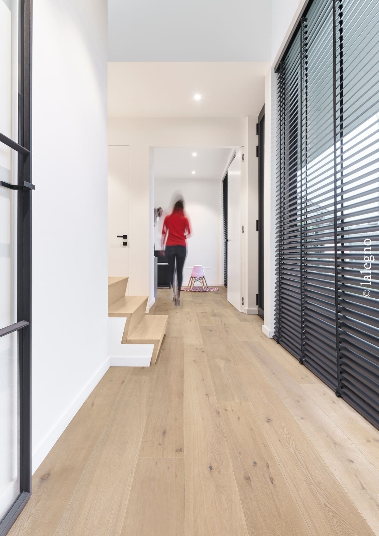 Couloir design avec parquet chêne clair et escalier moderne. Stores vénitiens sombres. Silhouette floue marchant vers une pièce lumineuse.