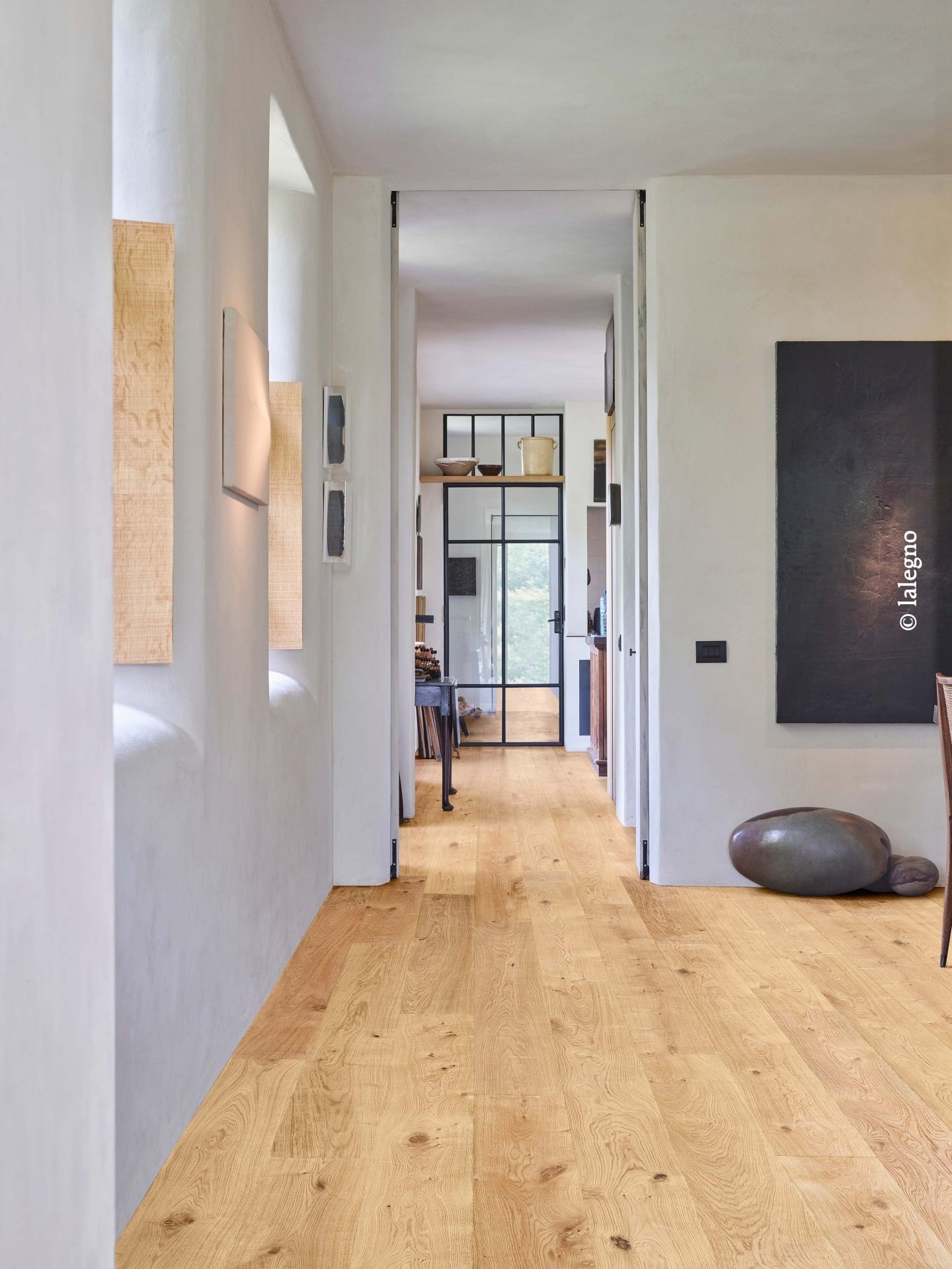 Parquet chêne clair dans couloir lumineux aux murs blancs. Porte vitrée noire, décor minimaliste et œuvre d'art noire.
