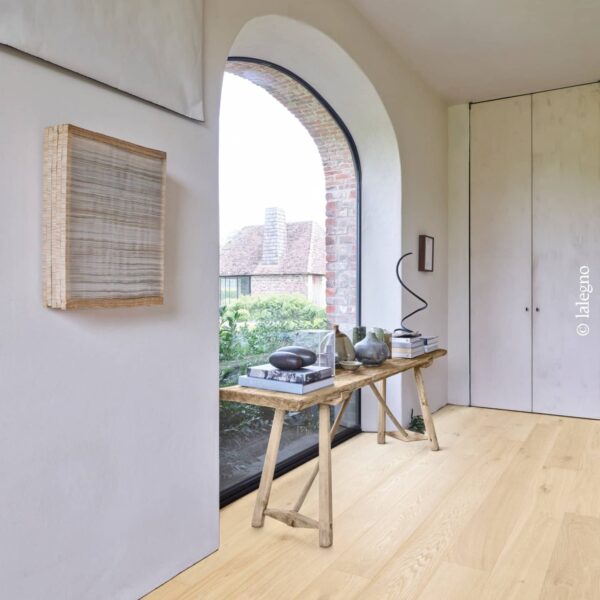 Entrée lumineuse avec large parquet en chêne clair Lalegno. Console rustique, œuvres d'art murales et fenêtre en arc. Style rustique chic.