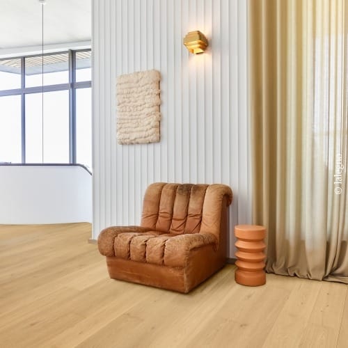 Fauteuil rouille confortable sur parquet chêne clair. Mur blanc flûté avec applique moderne dorée, rideaux beige et table terracotta.