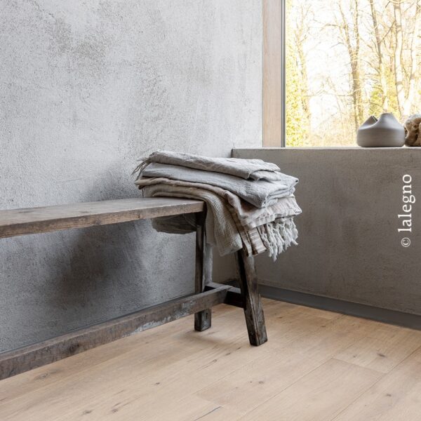 Parquet chêne clair dans un décor minimaliste : mur texturé gris, banc rustique en bois foncé, textiles lin près d'une fenêtre lumineuse.