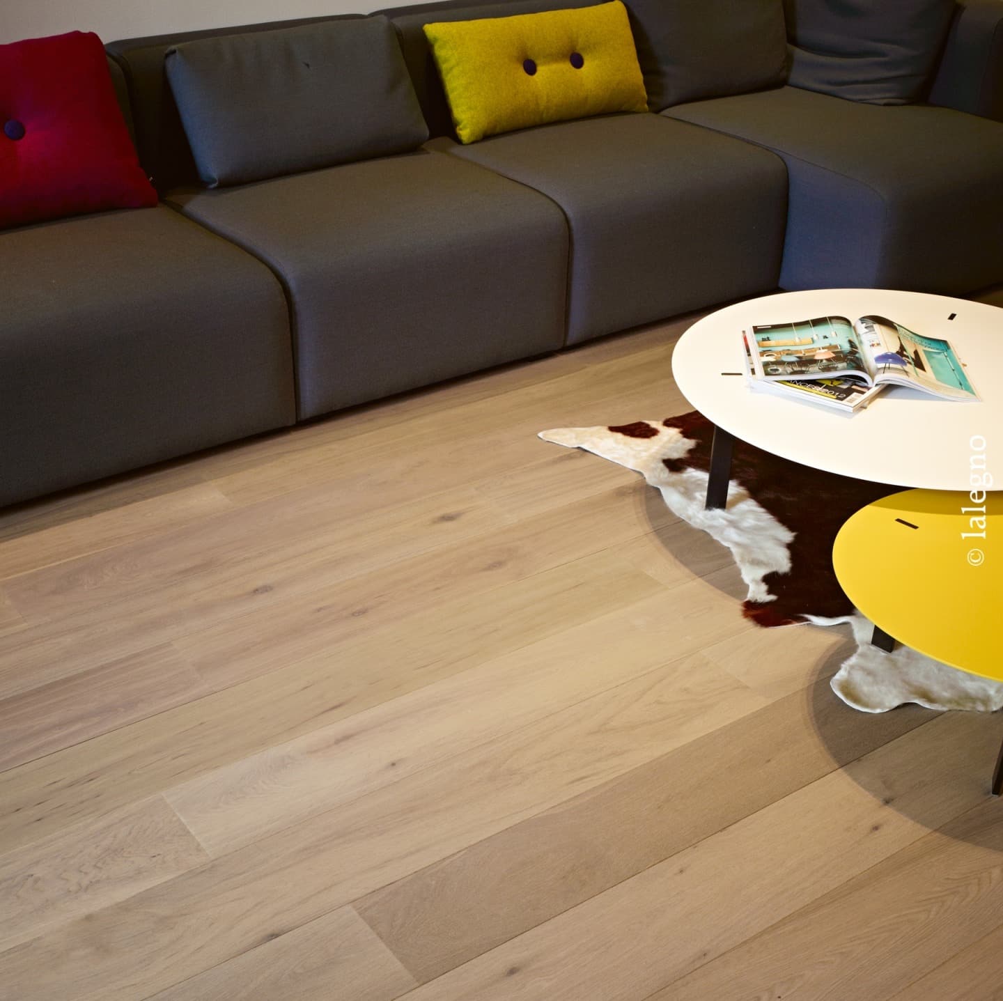 Parquet chêne clair à larges lames dans un salon moderne. Canapé gris, coussins rouge et jaune, table blanche et tapis peau de vache.