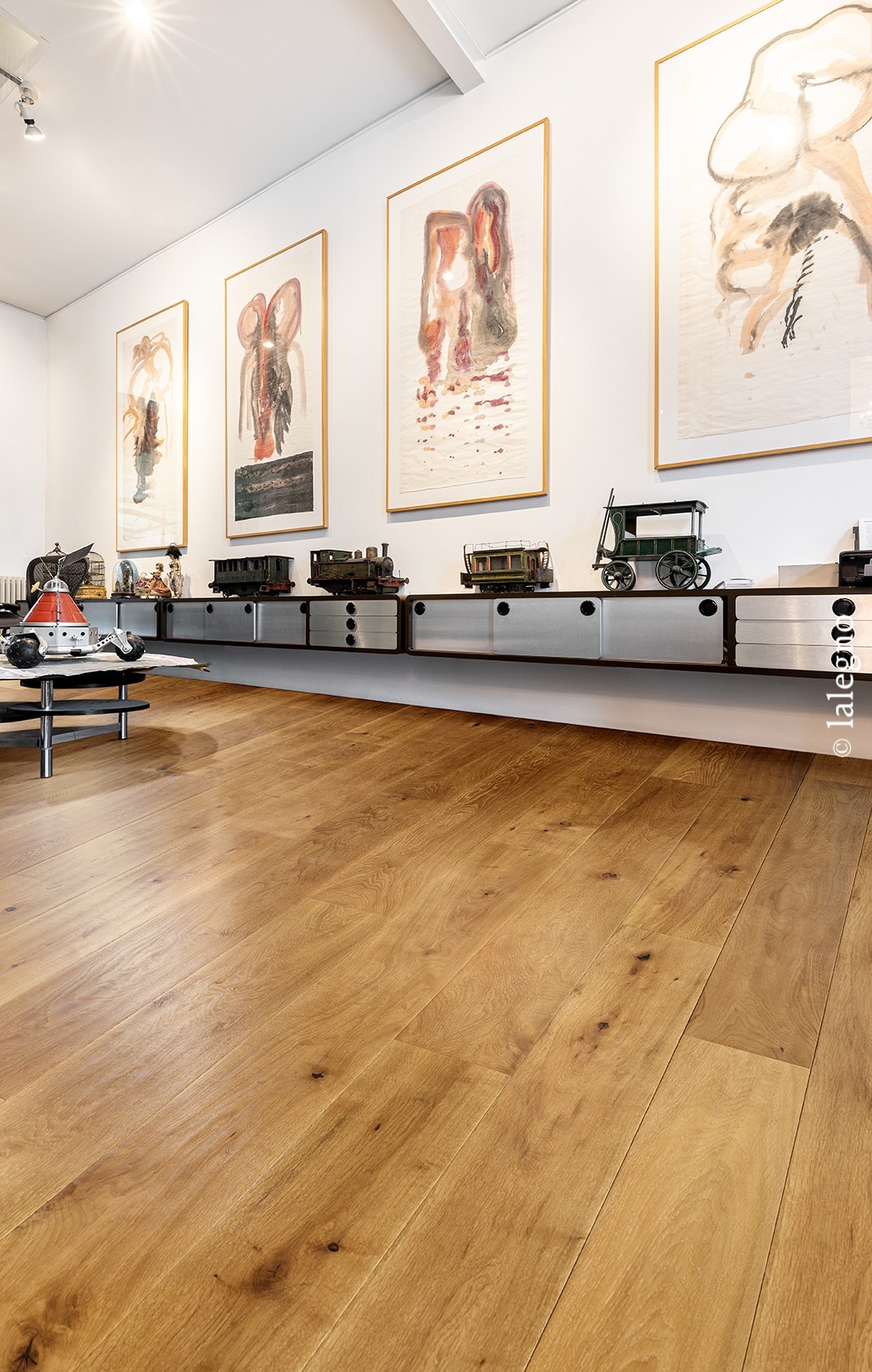 Parquet chêne doré en planches larges dans un intérieur moderne. Étagère flottante, jouets anciens, et grandes toiles abstraites encadrées.