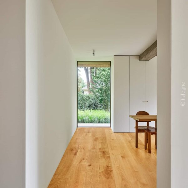 Couloir moderne minimaliste. Parquet chêne clair, murs blancs et plafond béton brut. Fenêtre panoramique donnant sur un jardin verdoyant.