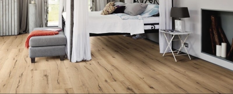 Parquet chêne naturel à planches larges, posé dans une chambre au style rustique-chic. Lit surélevé, pouf gris et cheminée décorative.