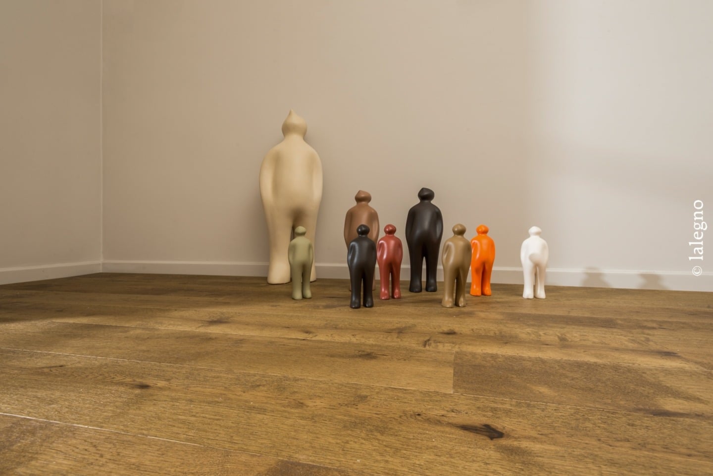 Collection de figurines décoratives colorées devant une sculpture géante beige, sur un sol bois rustique (parquet) (© lalegno).