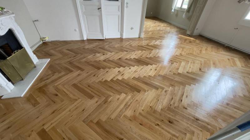 Parquet en chevrons de bois clair fraîchement vitrifié dans un salon rénové. Cheminée classique en marbre blanc et portes doubles.