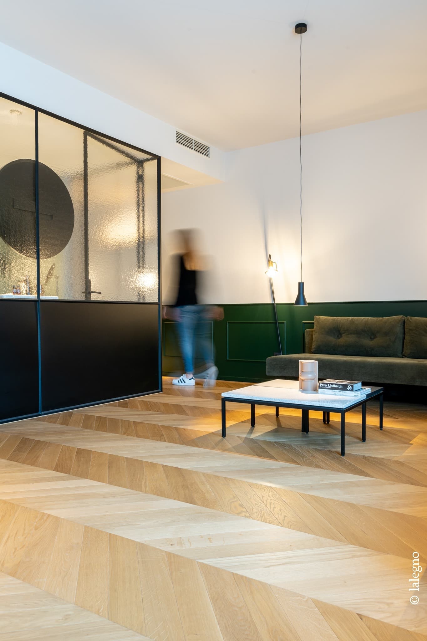 Parquet chêne chevron dynamique contrastant avec une cloison noire vitrée et un mur vert lambrissé. Peter Lindbergh sur la table.