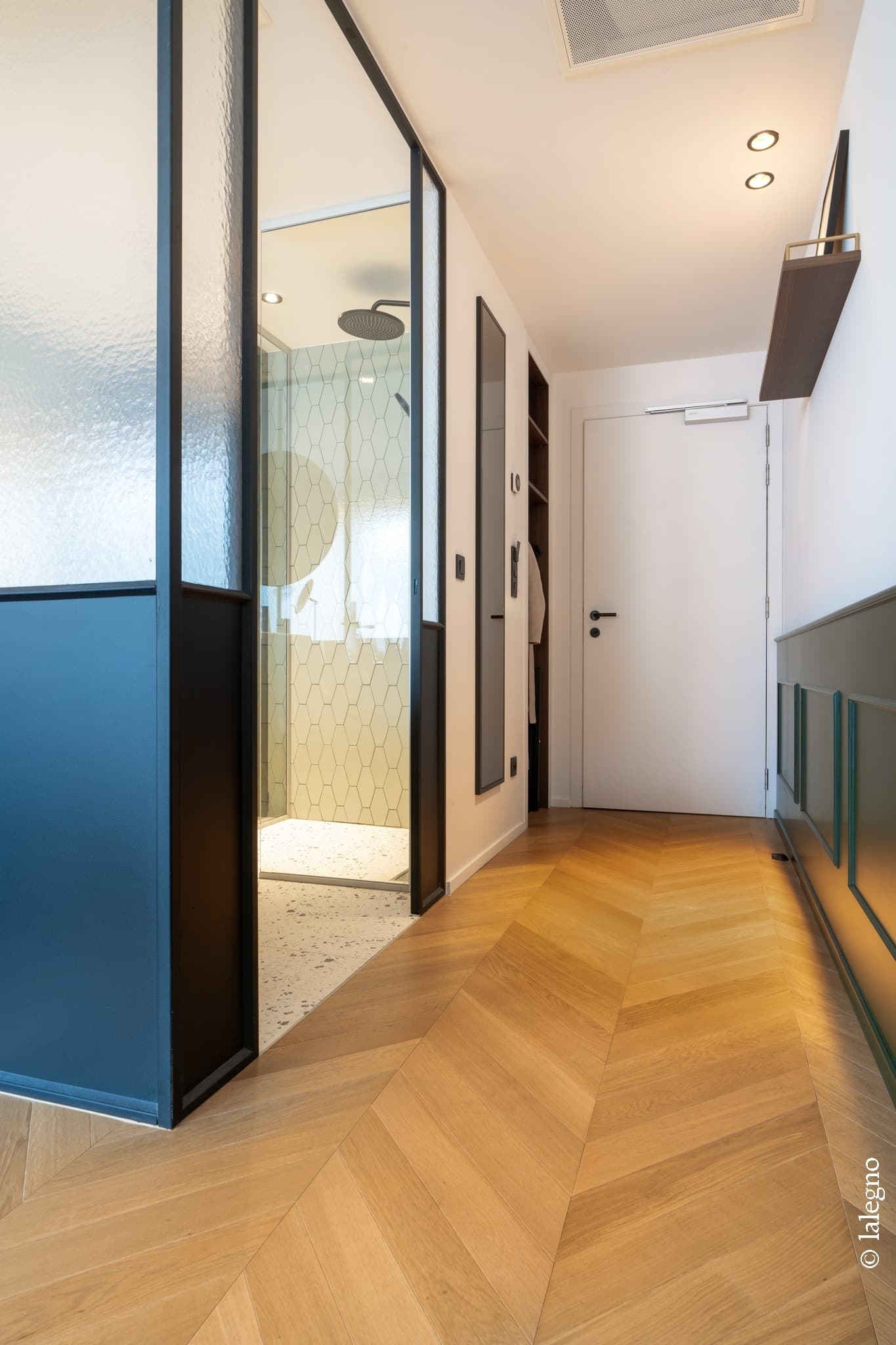 Couloir moderne avec parquet en chêne clair à chevrons. Verrière noire ouvrant sur une douche carrelée de hexagones et murs lambrissés vert foncé.