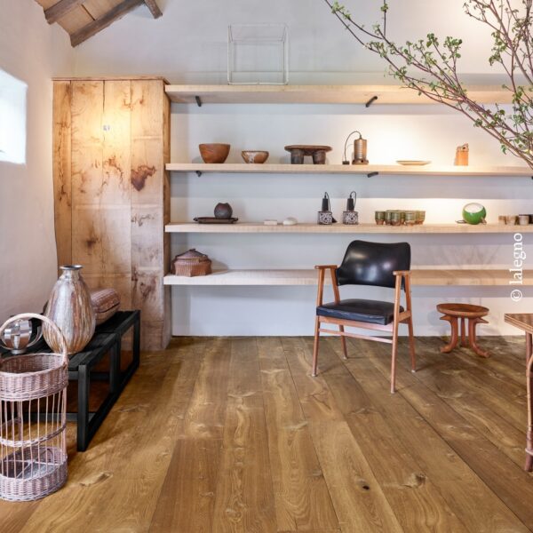 Plancher en chêne rustique à larges lames sous des poutres apparentes. Étagères flottantes minimalistes, fauteuil design et artisanat.