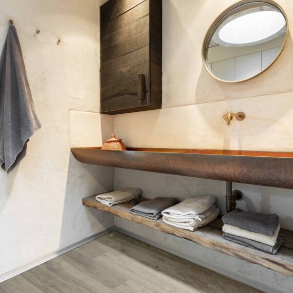 Salle de bain au design rustique industriel avec plancher rigide gris clair. Longue vasque en cuivre, miroir rond et armoire murale foncée.