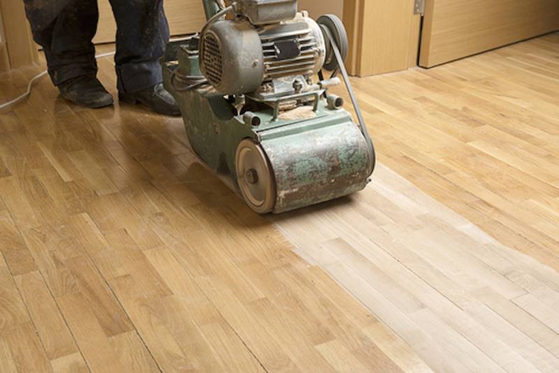 Ponceuse professionnelle décape un parquet en bois. Le ponçage retire la finition, exposant les lattes de bois brut, clair et rénové.