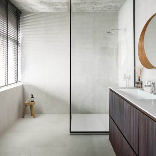 Salle de bain moderne avec carrelage style béton gris clair et plafond brut. Douche vitrée, vanité en bois foncé et miroir rond.