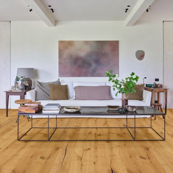 Pièce de vie épurée : murs blancs, canapé, longue table basse en métal. Éclairage directionnel et sol en parquet chêne rustique.